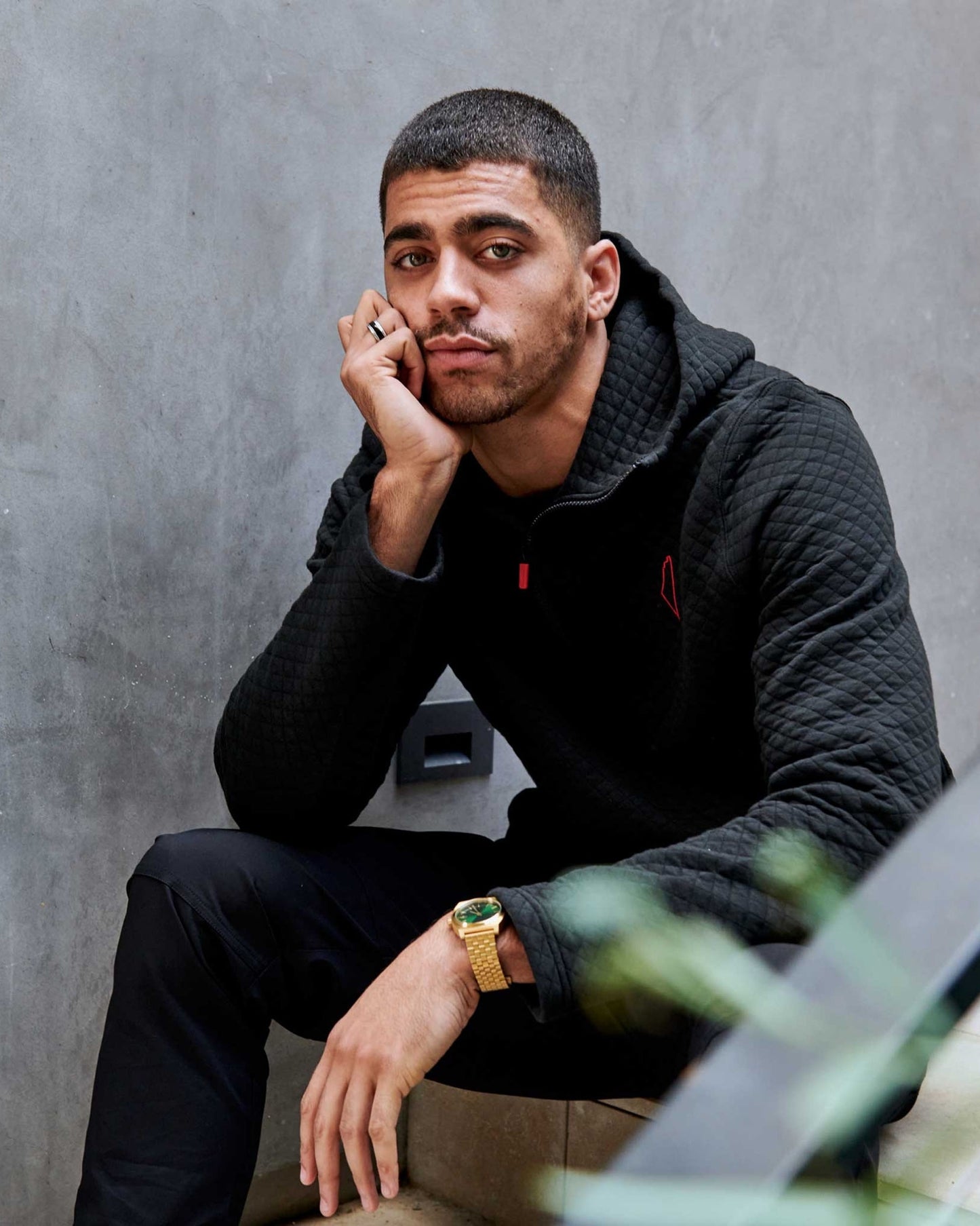 A young man with short hair in a black textured hoodie with red details, dark trousers and gold watch, sits against a gray wall, resting his chin on his hand, looking contemplative.