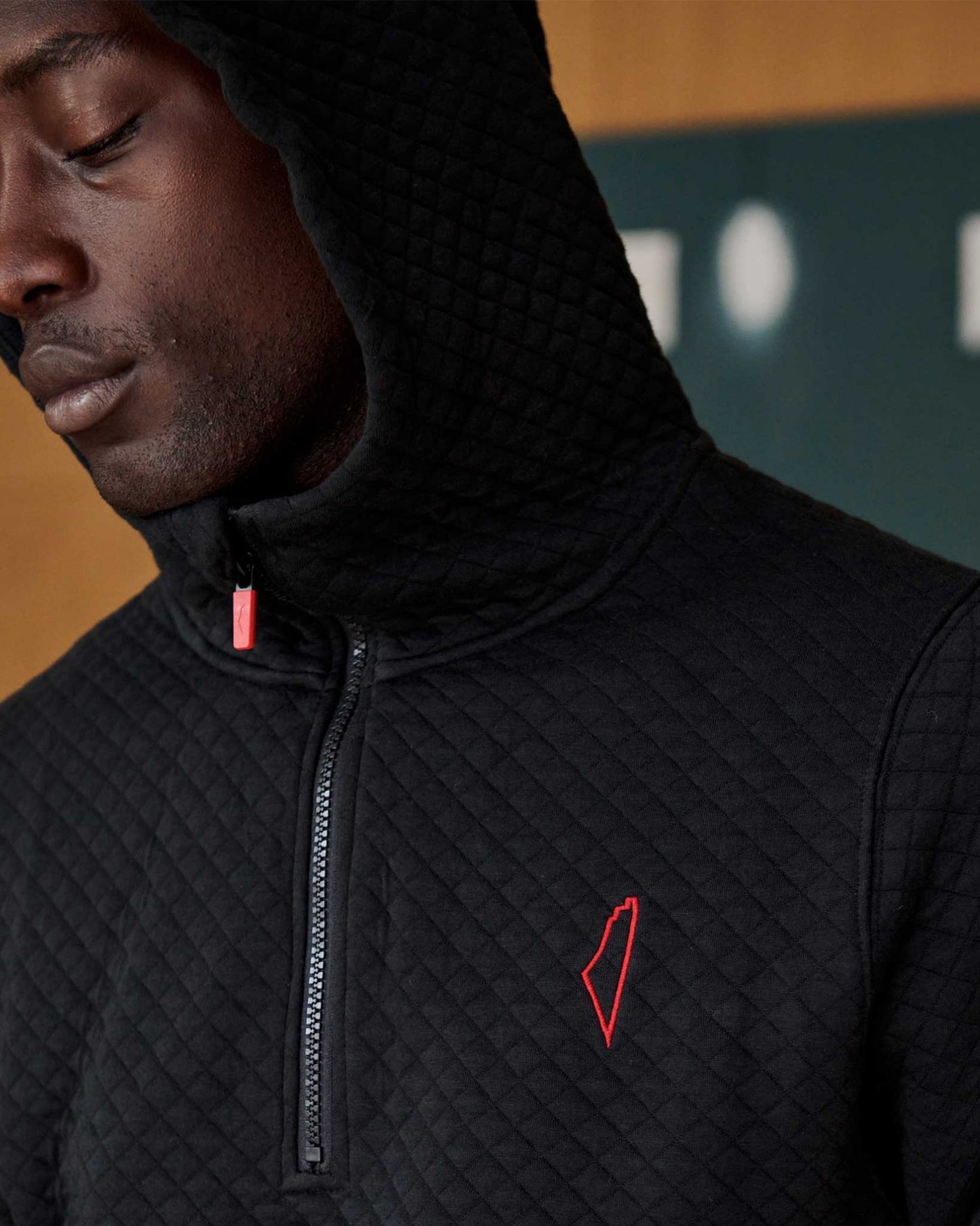 Close up of a person wearing a black quilted hoodie with a red stitched logo on the left chest. The hoodie is partially zipped.