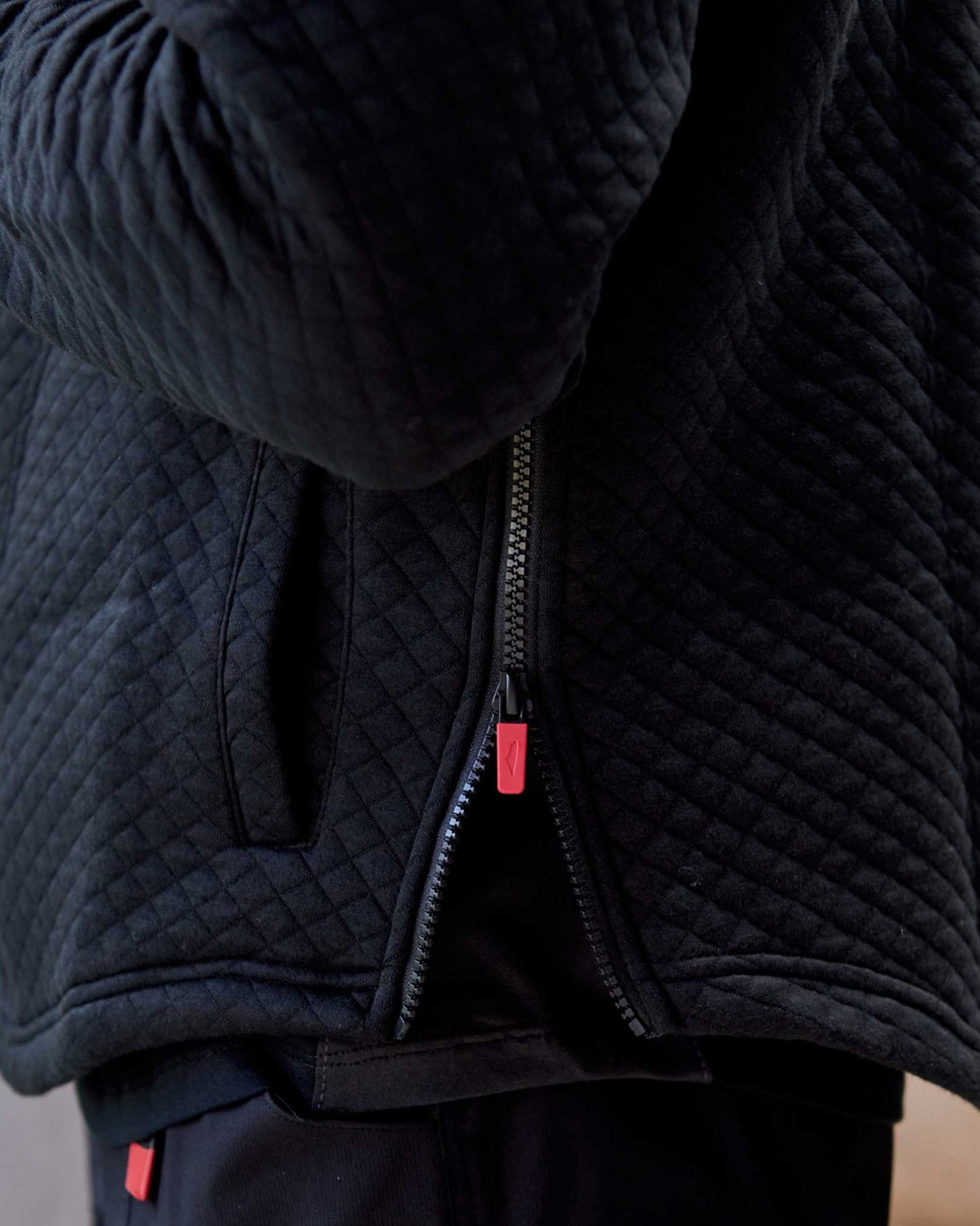 Close-up of a black hoodie with a partially opened zipper, featuring red pull tabs. The texture appears cozy and stylish.