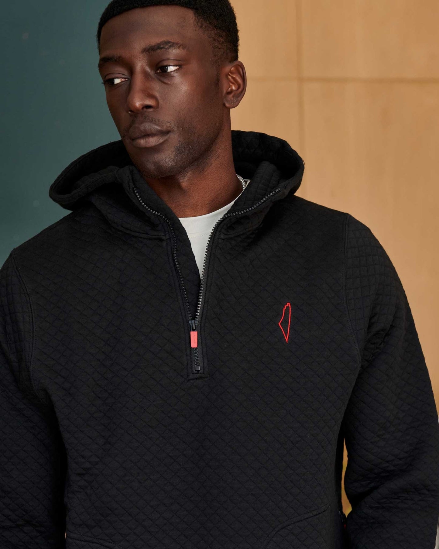 A man wearing a black quilted hoodie with a red stitched logo on the left chest, over a white top. The hoodie is partially zipped.