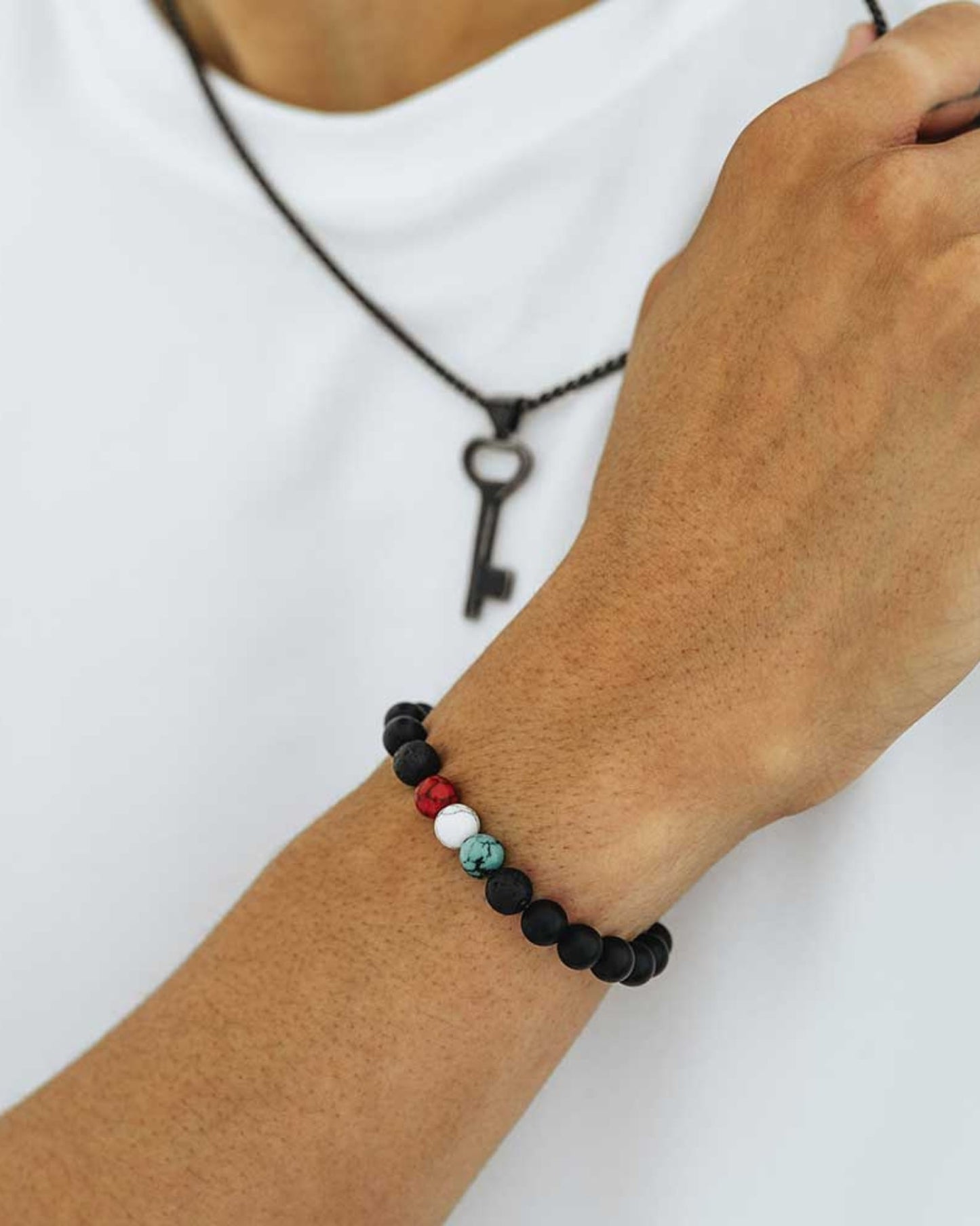 A close-up of a man's wrist adorned with a bracelet in tones of black, white, green and red, wearing a white top and a key pendant.