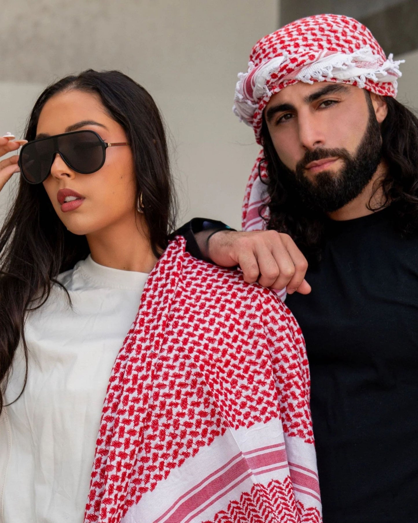 A man and woman wearing sunglasses are dressed in traditional Arabic clothing, posing with a white and red Kufiya, standing together in a culturally rich setting.