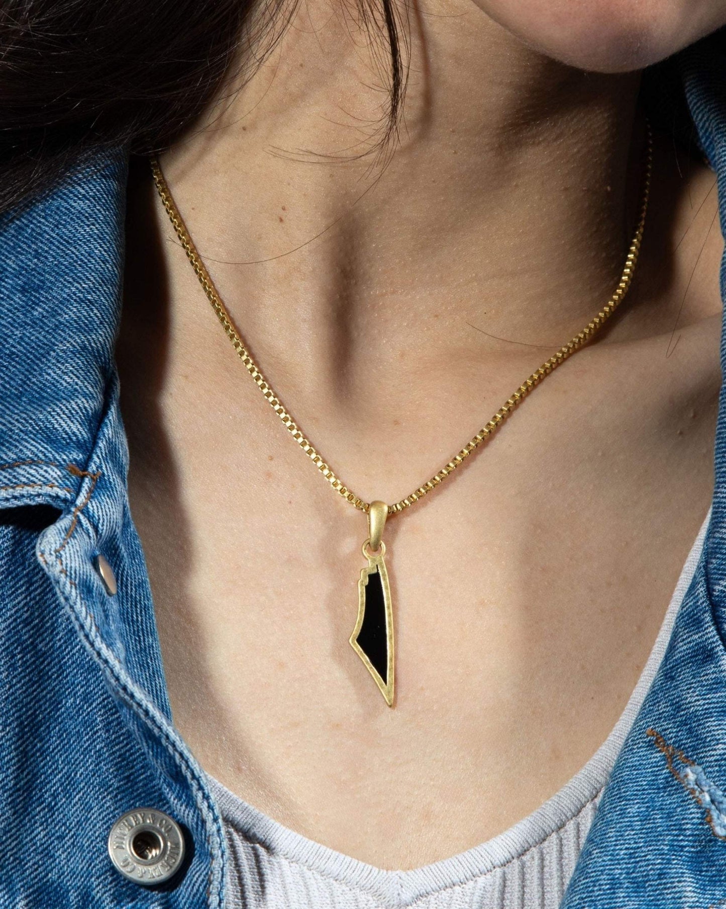 A close-up of a woman with a denim jacket displaying a gold necklace on her neck, highlighted by a distinctive black stone pendant.