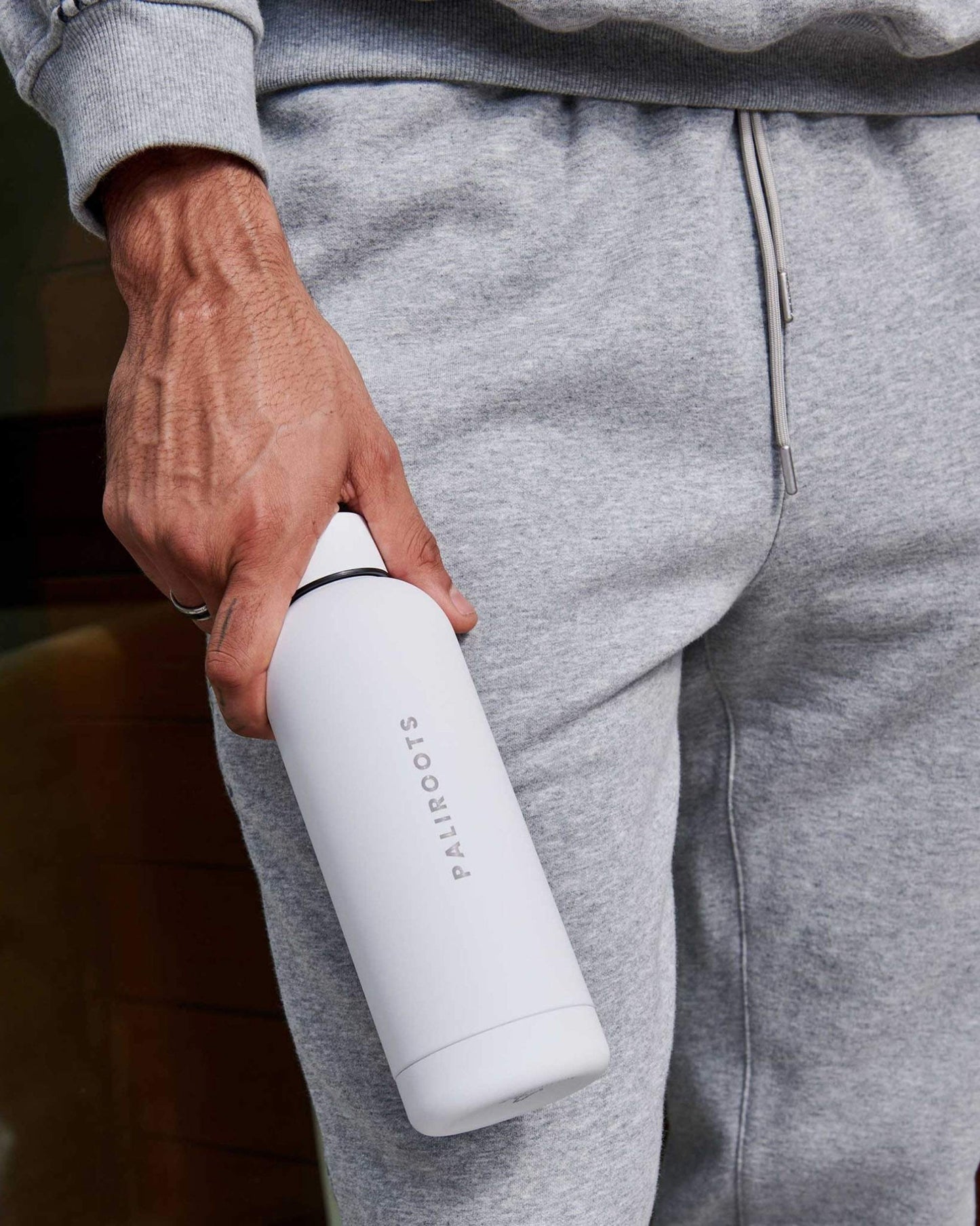 A person in a grey tracksuit holds a white water bottle, reading "Paliroots" in its center, suggesting a sporty or casual lifestyle.