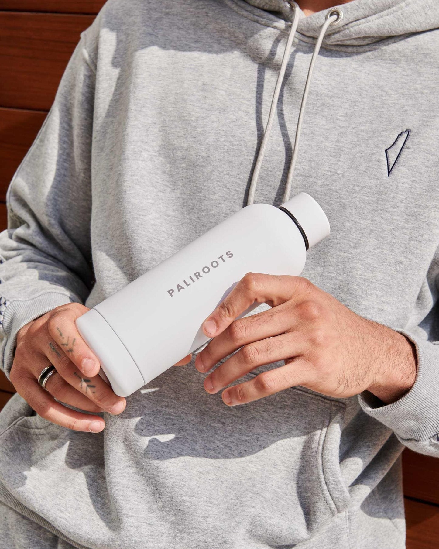 A person in a grey tracksuit holds a white water bottle, reading "Paliroots" in its center, suggesting a sporty or casual lifestyle.