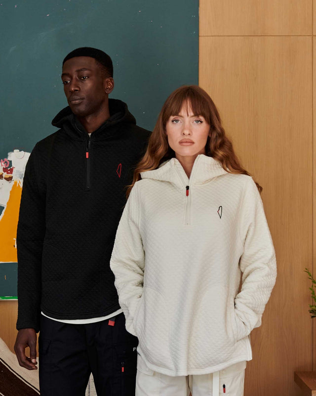 Two people stand in a room wearing quilted pullovers, one black with red details, and the other white with multicoloured details, against a backdrop of teal and wood panel walls.